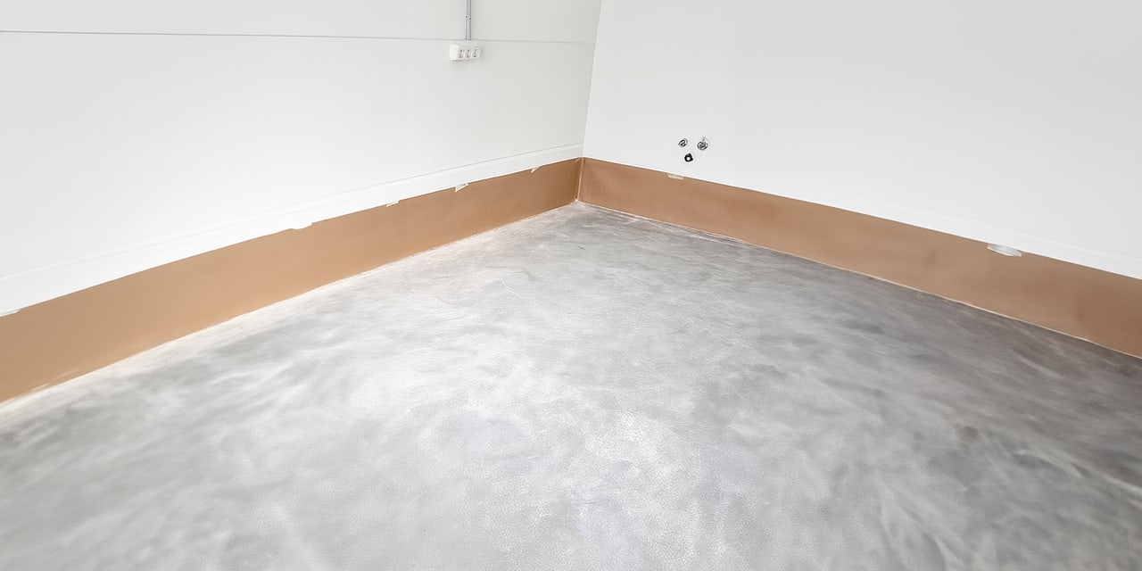 Geschädigte Bodenecke mit grauem Boden, braunem Sockelschutz und weißen Wänden vor der Bodenbeschichtung